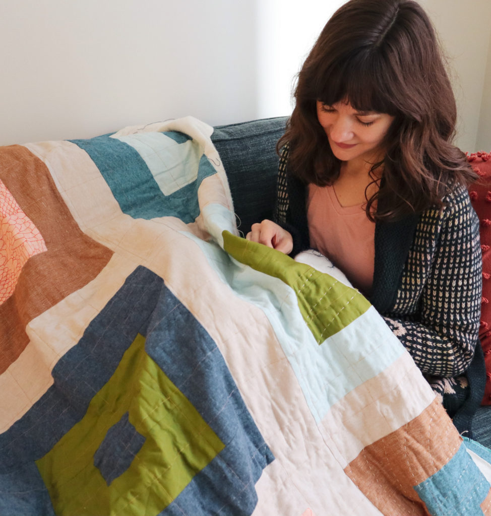 (Very) Slow Stitching 3 Meredith wears a pink shirt and blue cargidan and sits on her couch, with a quilt spread over her that she hand quilts happily