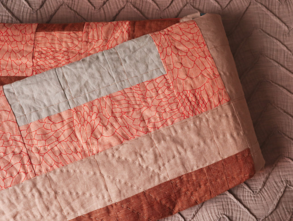 (Very) Slow Stitching 2 a beige, coral, blue and brown pieced quilt with hand quilting in progress on a grey duvet cover