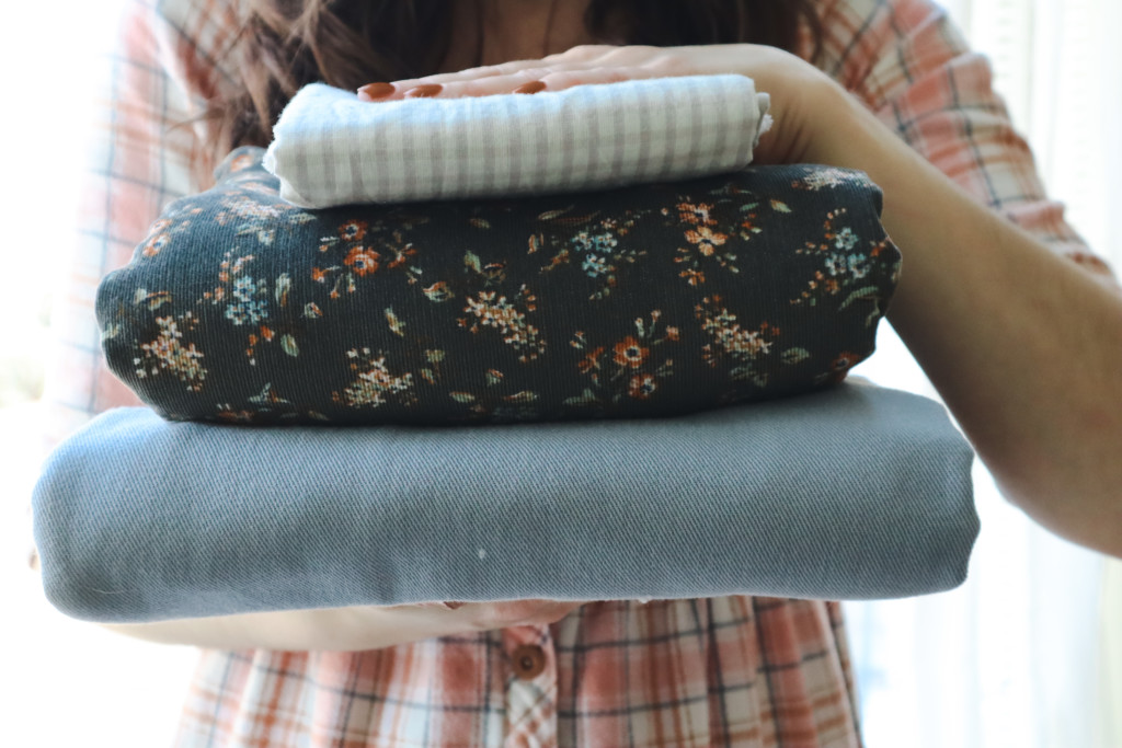 Meredith holds a stack of twill, corduroy and woven cotton fabric in front of her check while wearing a flannel hinterland dress
