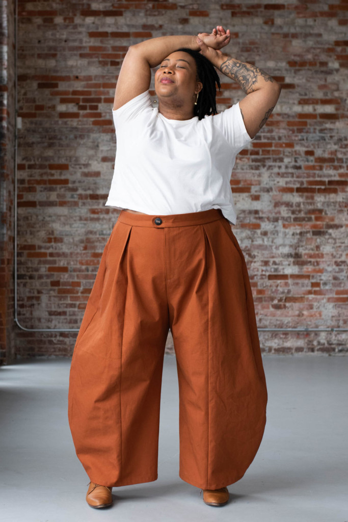 Ashley wears orange Arthur Pants and a white tee in front of a brick wall..