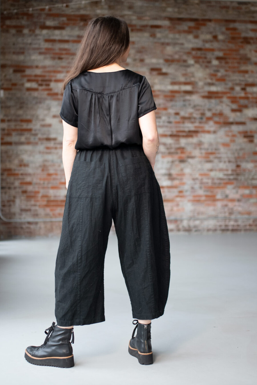 Meg is wearing a black Nocturne Top and black Arthur Pants. She is turned away from the camera in front of a brick wall.