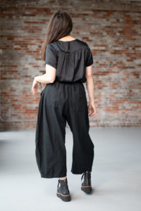 Meg is wearing a black Nocturne Top and black Arthur Pants. She is turned away from the camera in front of a brick wall.