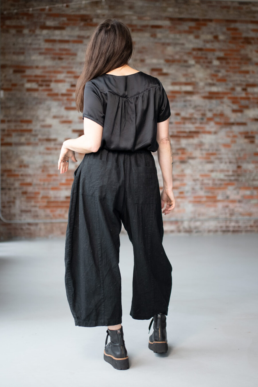 Meg is wearing a black Nocturne Top and black Arthur Pants. She is turned away from the camera in front of a brick wall.