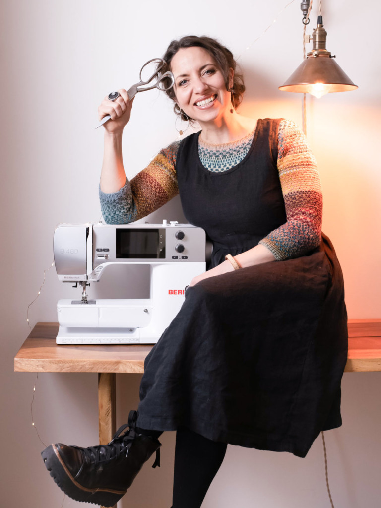 Learn to Fit and Sew Bodices 1 Meg wears a black, sleeveless dress over a colorful handknit sweater. She is sitting next to her sewing machine and holding scissors in her hand.