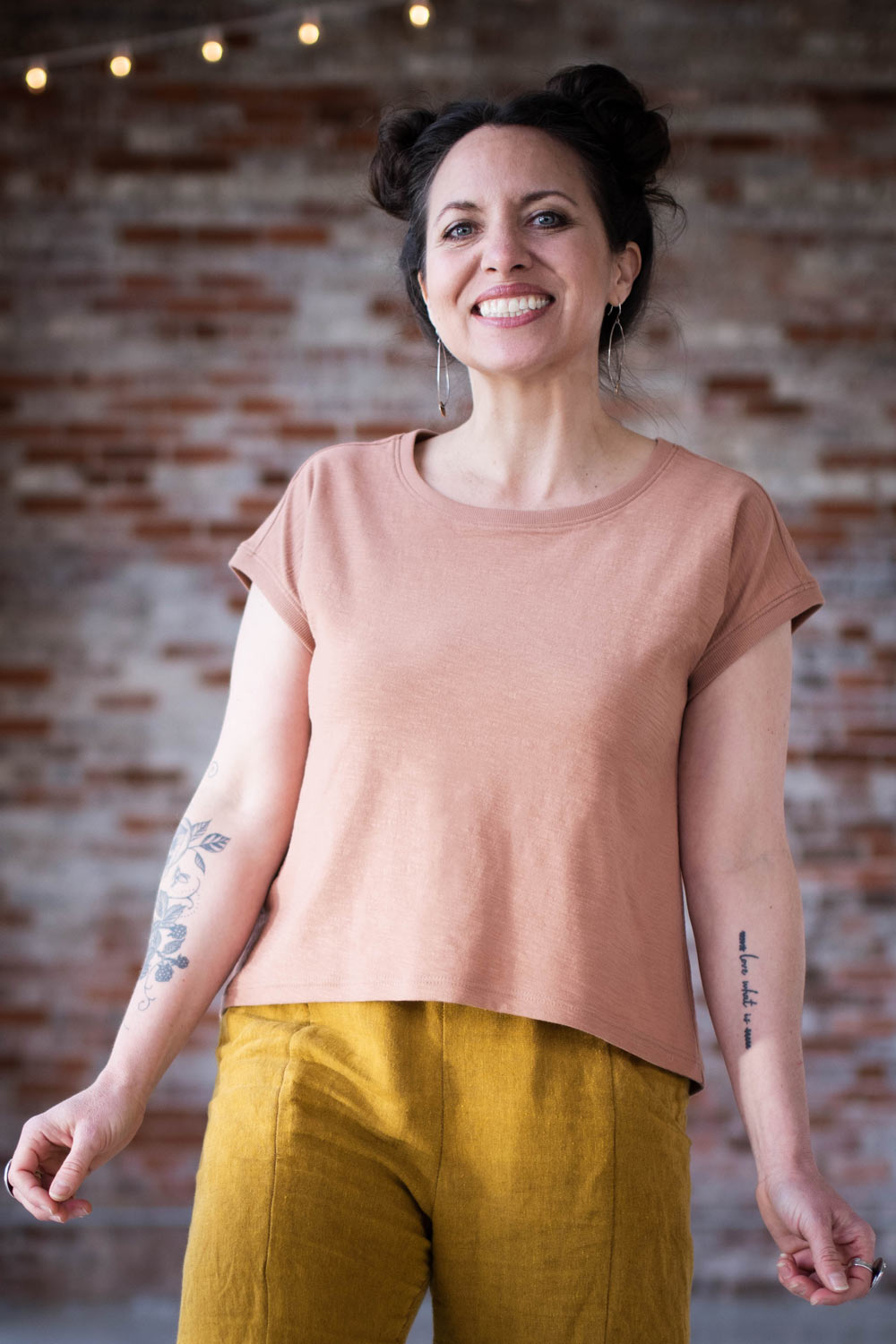 Meg wears a pink bedrock tee and yellow arenite pants, staring in front of a brick wall